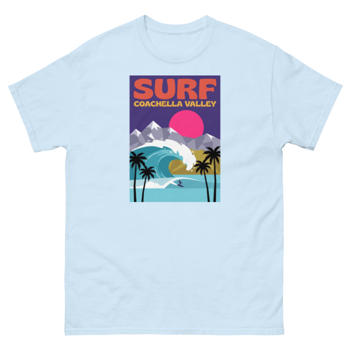 A light blue crewneck t-shirt featuring a colorful retro-style graphic print on the chest that reads 'SURF COACHELLA VALLEY' above an illustration of a surfer, palm trees, mountains, and a pink sun.