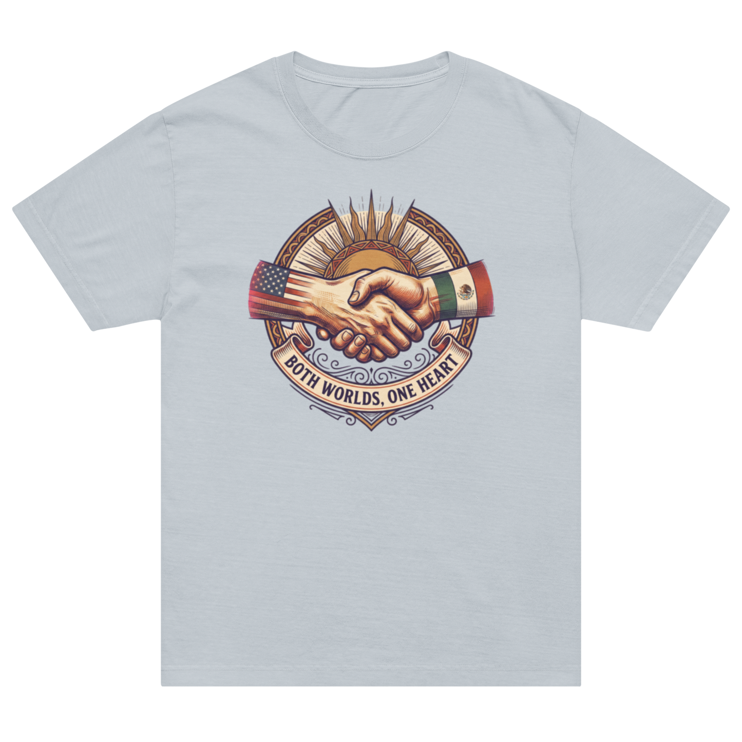 A light blue-gray short-sleeve crewneck t-shirt featuring a central graphic of two hands shaking, with one hand wearing a US flag sleeve and the other wearing an Mexican flag sleeve, under a sunburst and above the text 'BOTH WORLDS, ONE HEART'.