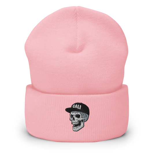 A light pink knit beanie hat with a folded cuff, featuring an embroidered graphic of a gray and black skull wearing a black cap with the word 'CALI' in white.