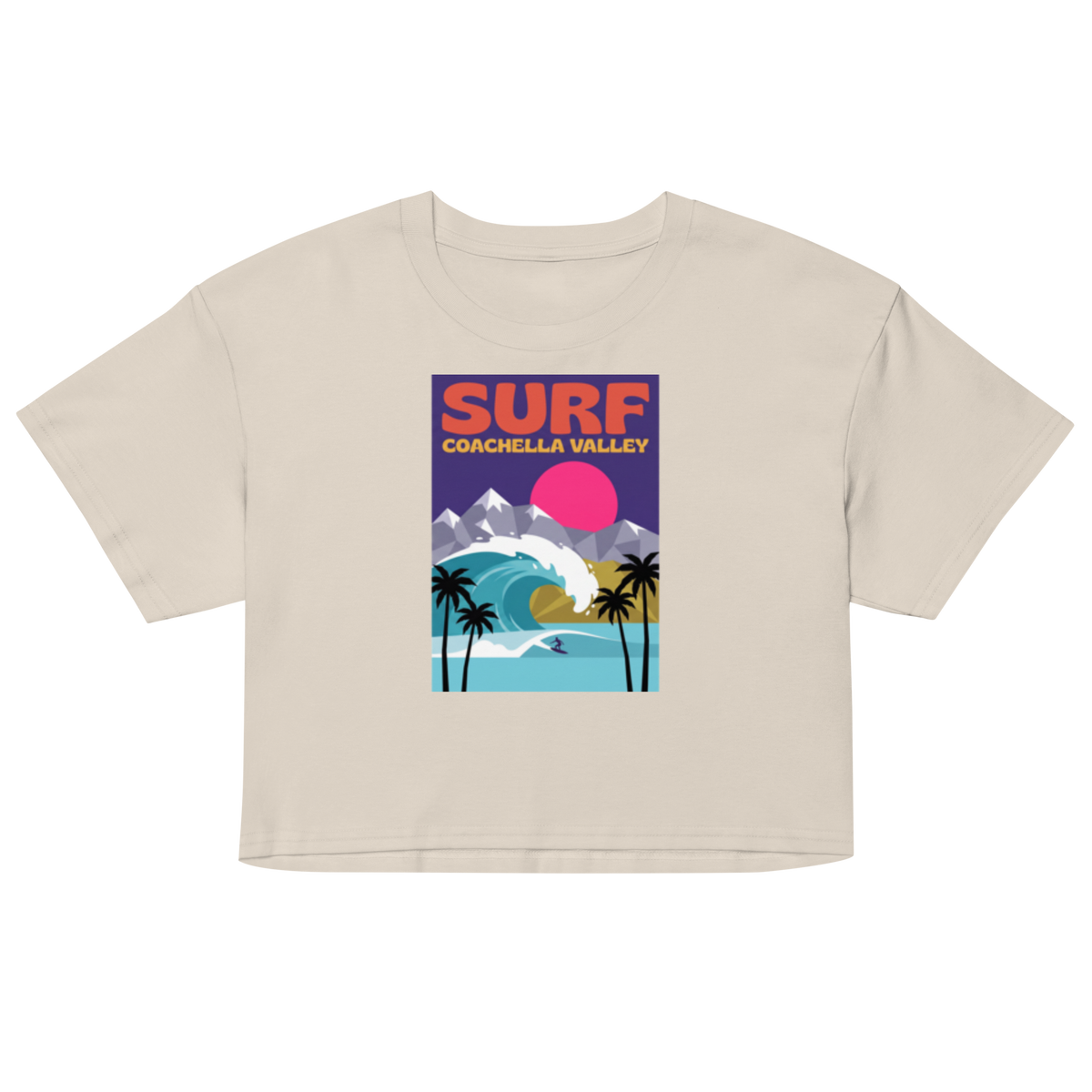 A light beige short-sleeve crop top featuring a colorful retro-style graphic print on the front with the text 'SURF COACHELLA VALLEY' and an illustration of a wave, mountains, palm trees, and a pink sun.