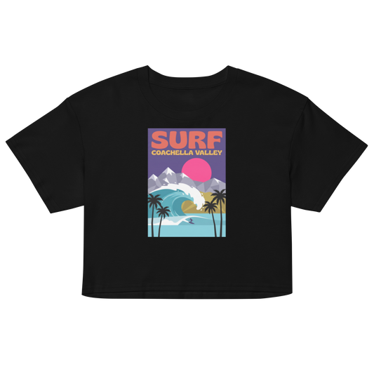 A black short-sleeve crop top t-shirt featuring a colorful retro graphic on the chest that reads 'SURF COACHELLA VALLEY' above an illustration of palm trees, a wave, mountains, and a pink sun.