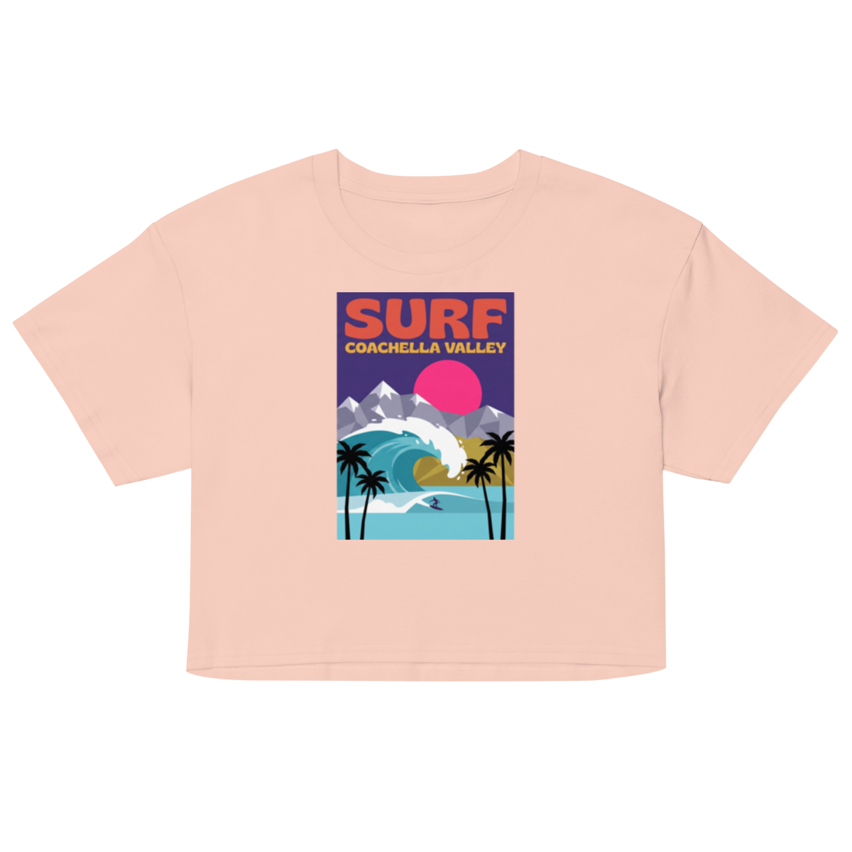 A light peach-pink short-sleeve crop top featuring a colorful retro-style graphic on the chest that reads 'SURF COACHELLA VALLEY' above an illustration of palm trees, mountains, a wave, and a pink sun.