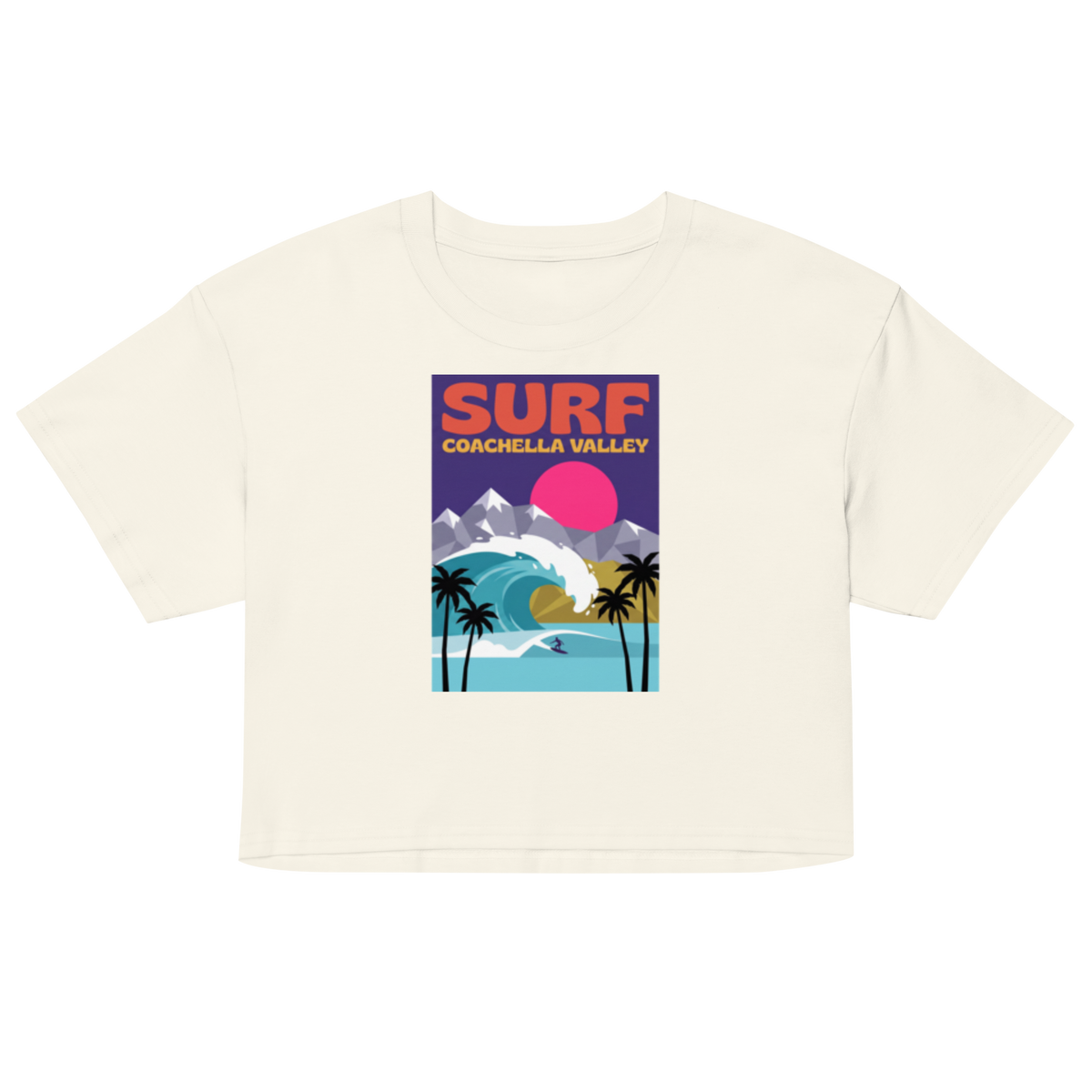 An off-white short-sleeve crop top featuring a colorful retro-style graphic print on the front with the text 'SURF COACHELLA VALLEY' and an illustration of a wave, mountains, palm trees, and a pink sun.