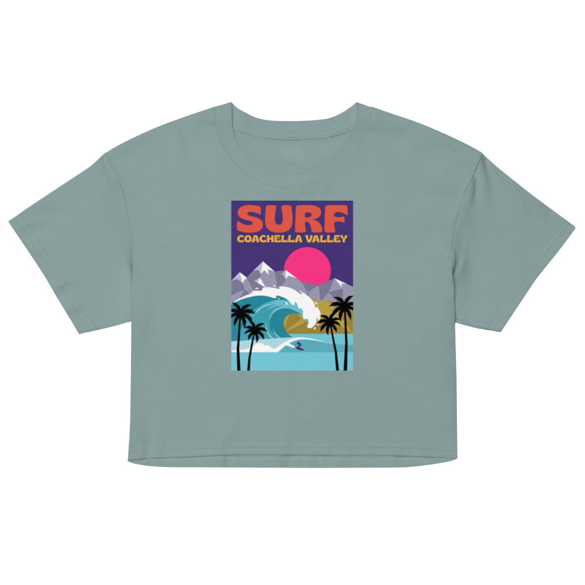 A sage green short-sleeve crop top featuring a colorful retro-style graphic on the chest that reads 'SURF COACHELLA VALLEY' above an illustration of mountains, a wave, palm trees, and a pink sun.