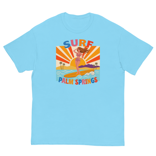 A light blue crewneck t-shirt featuring a retro-style graphic of a woman surfing on a yellow board with palm trees and the text 'SURF PALM SPRINGS' in colorful letters.