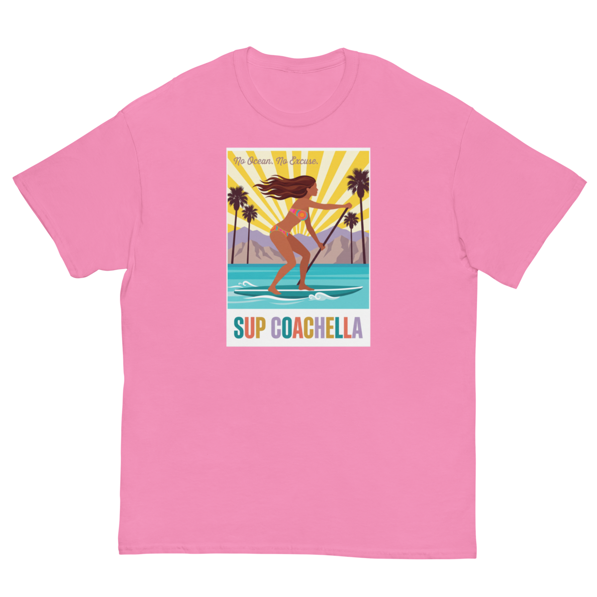 A pink short-sleeve crewneck t-shirt featuring a colorful graphic print on the front. The graphic shows a woman paddleboarding on turquoise water with palm trees and a yellow sunburst background, with the text 'My Dream Surf Spot' and 'SUP COACHELLA' below.