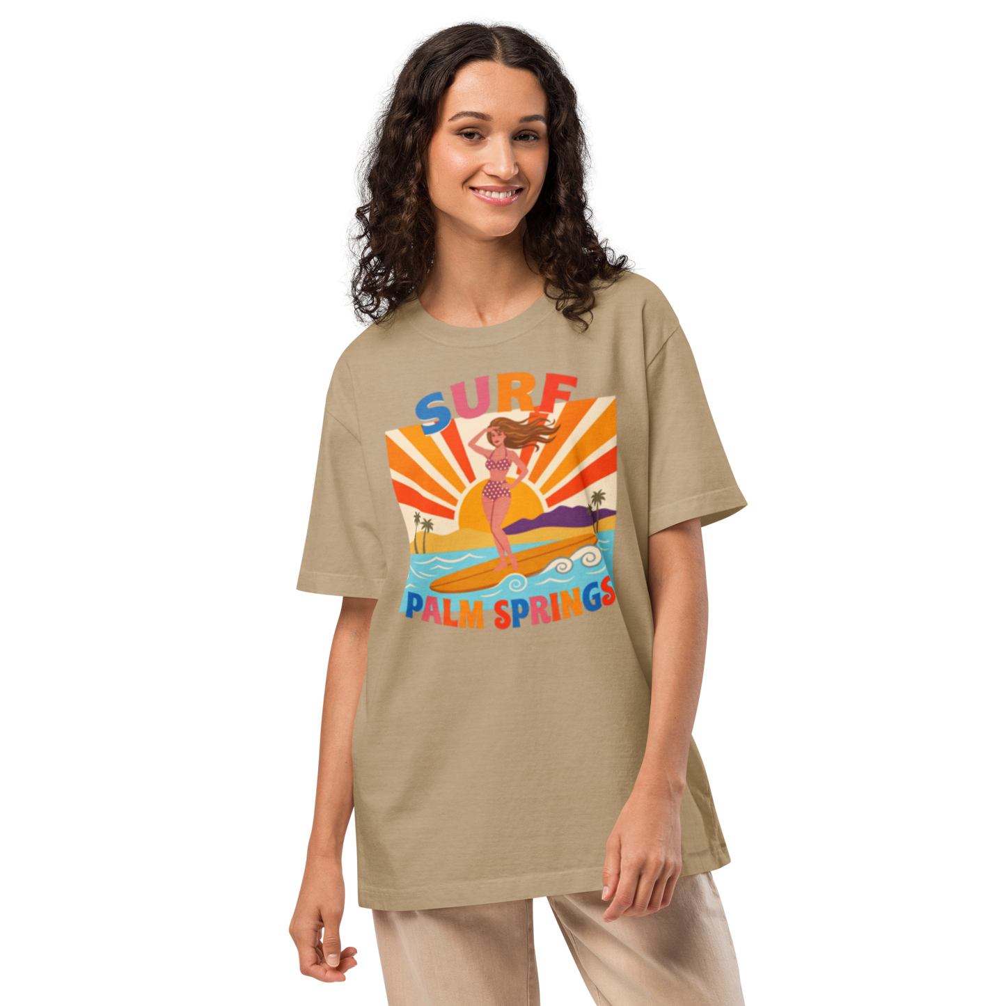 A woman modeling an oversized beige graphic t-shirt with a colorful 'Surf Palm Springs' design on the front, paired with light beige pants, against a plain white background.
