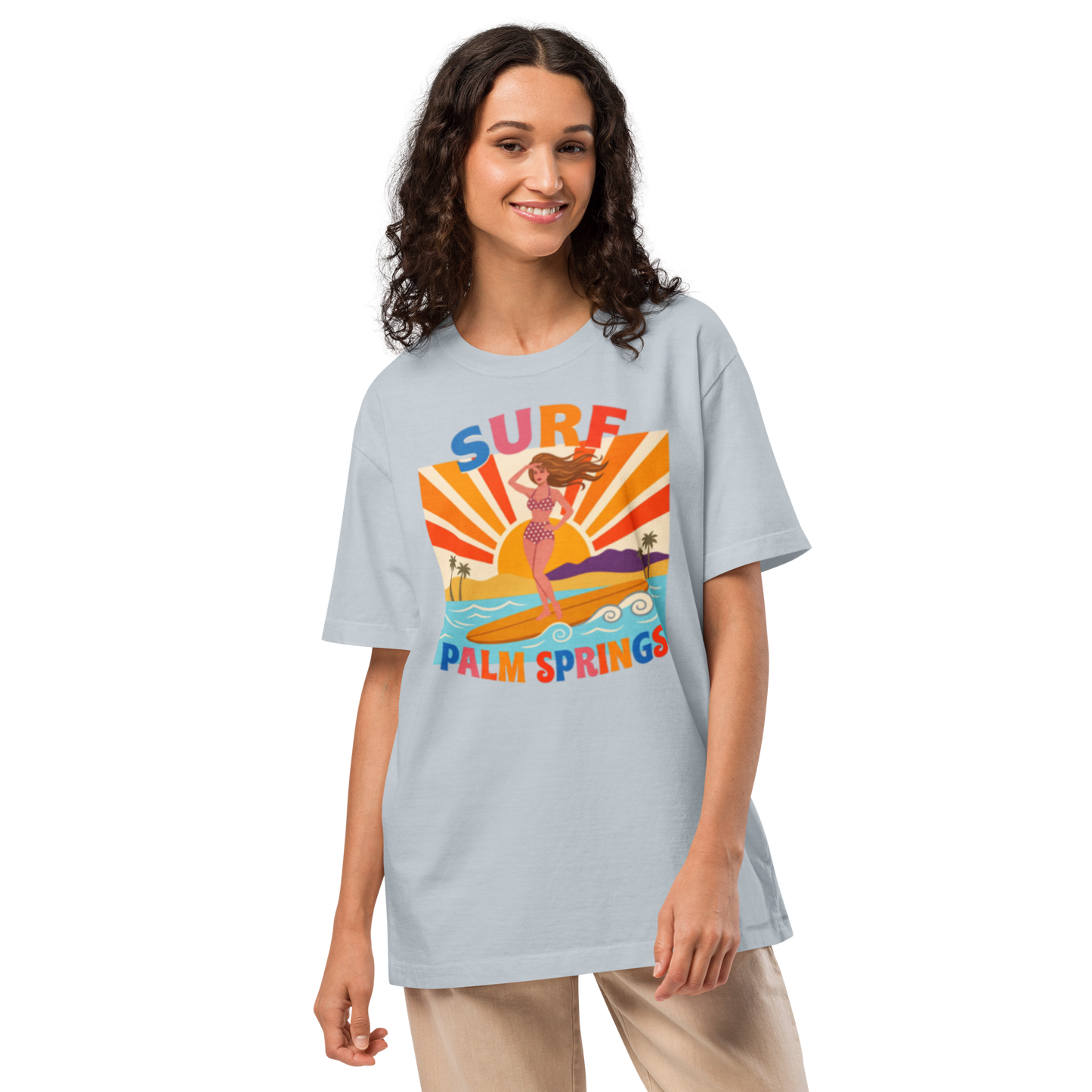 A woman modeling a light blue t-shirt with a colorful retro graphic that reads 'SURF PALM SPRINGS' featuring a mermaid and sun rays, set against a plain white background.