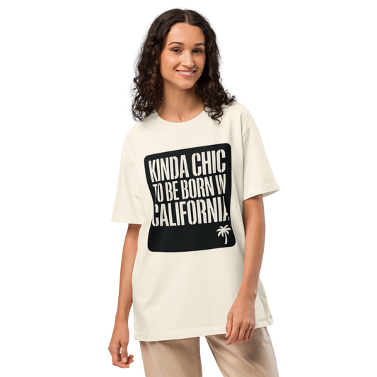 A woman modeling an oversized cream-colored t-shirt with a black graphic box on the front that reads 'KINDA CHIC TO BE BORN IN CALIFORNIA!' in white text, paired with beige pants, against a solid white background.