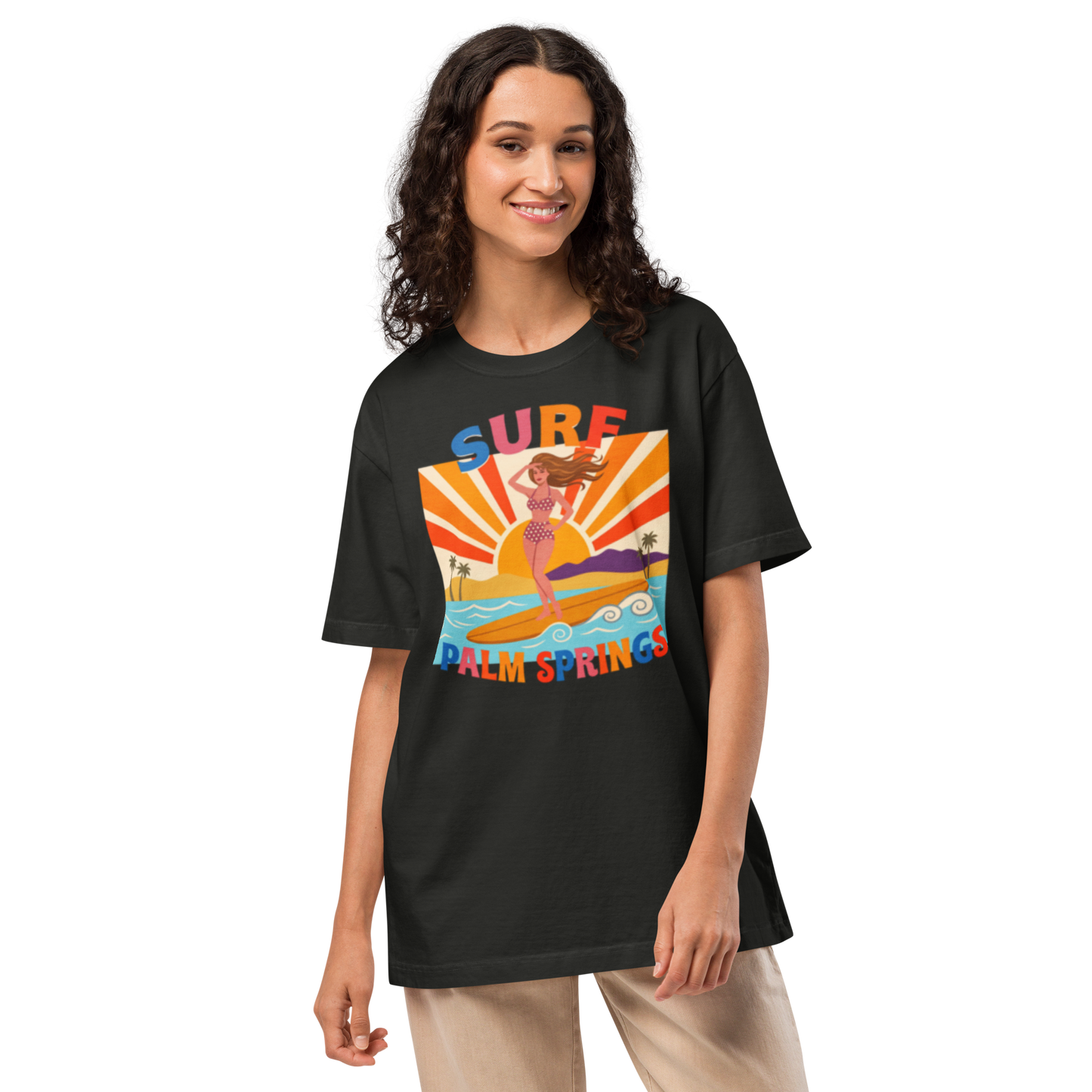 A woman modeling a black graphic t-shirt with a colorful retro-style 'Surf Palm Springs' design on the front, paired with beige pants, against a plain white background.