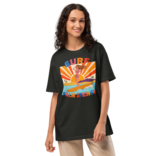 A woman modeling a black graphic t-shirt with a colorful retro-style 'Surf Palm Springs' design on the front, paired with beige pants, against a plain white background.