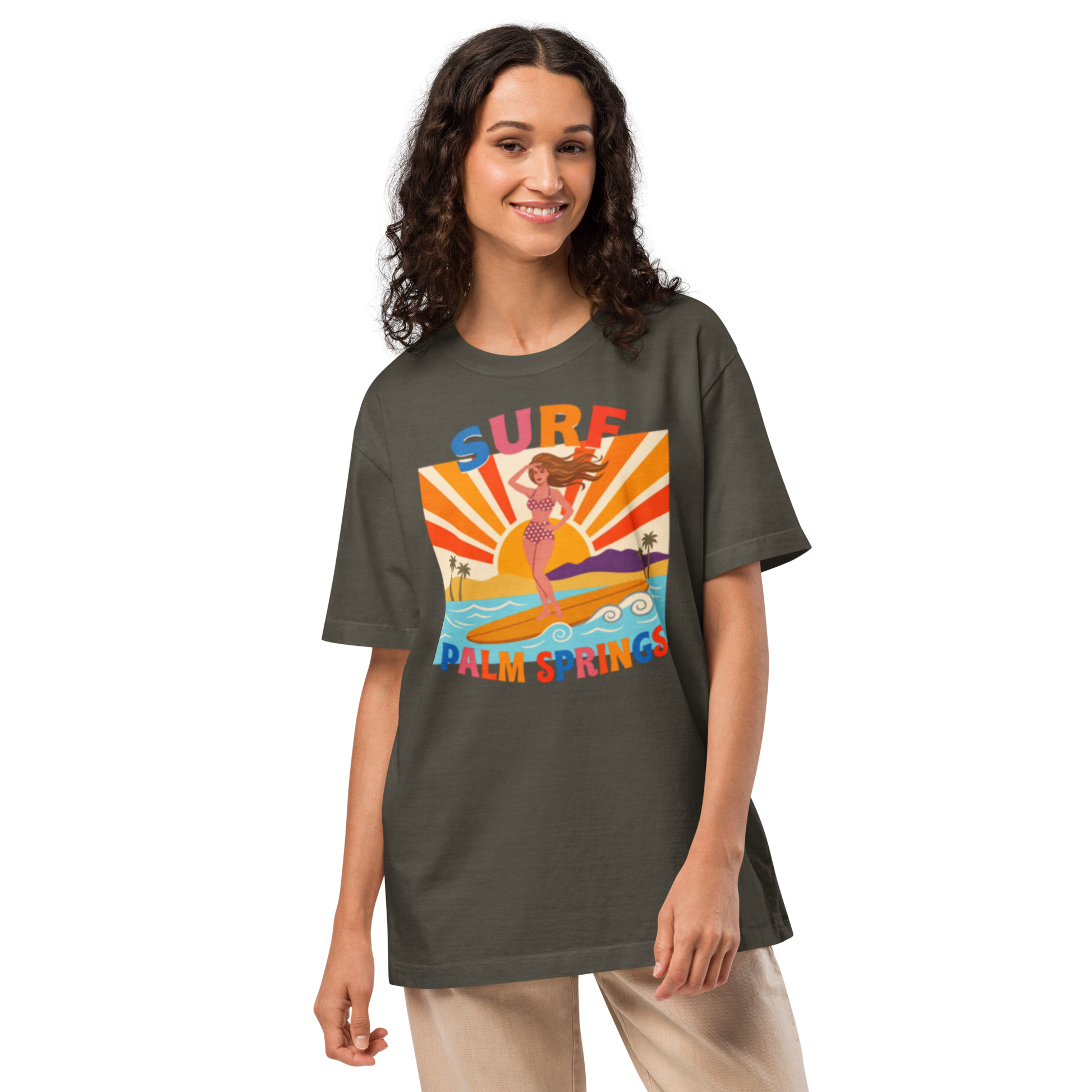 A woman modeling an oversized dark olive green t-shirt with a colorful retro graphic of a mermaid on a surfboard with the text 'SURF PALM SPRINGS'.