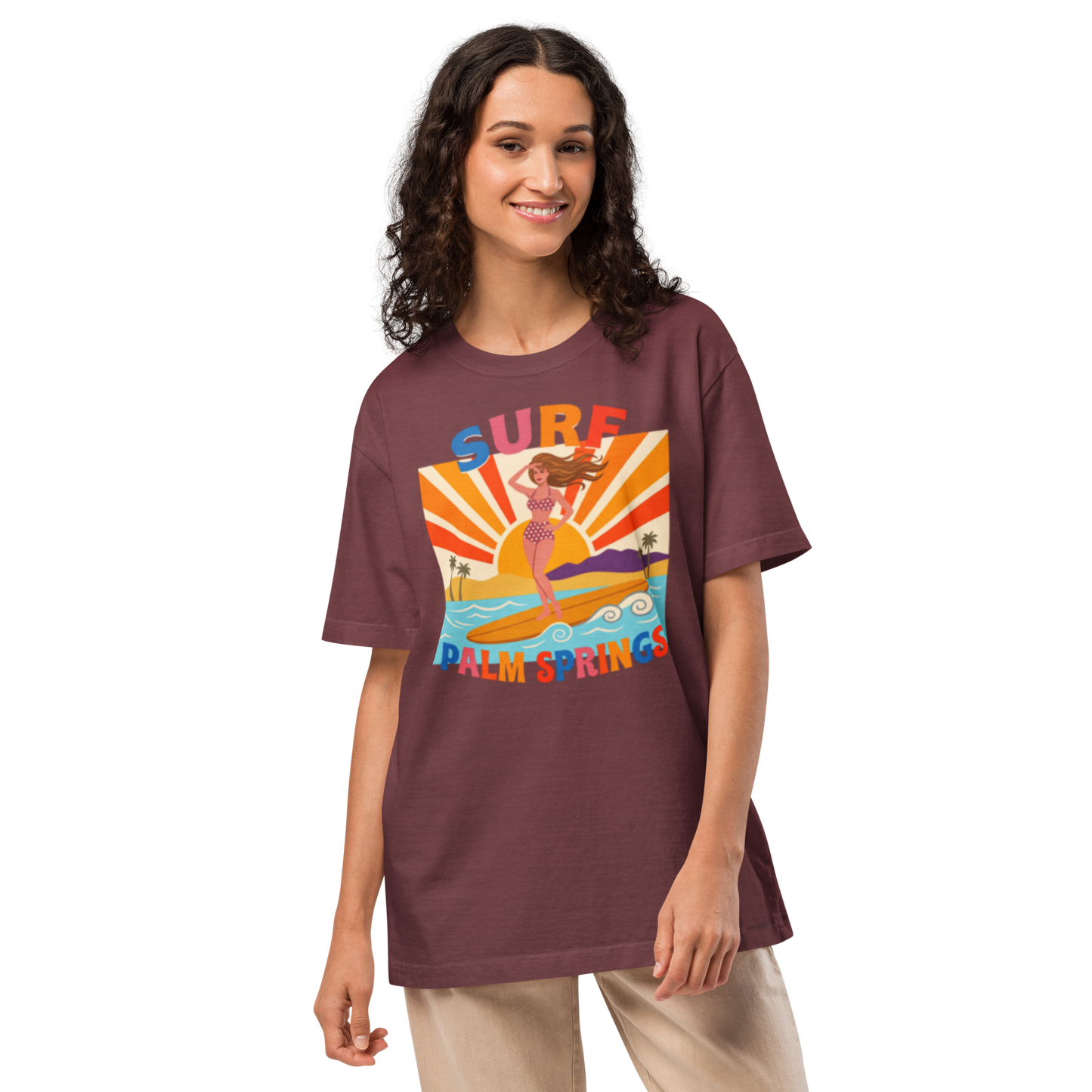 A woman modeling an oversized short-sleeve t-shirt in a dusty plum-brown color, featuring a colorful retro graphic of a mermaid on a surfboard with the text 'SURF PALM SPRINGS'.