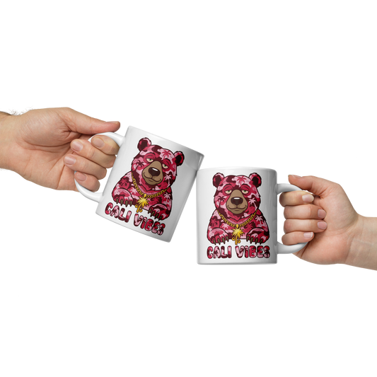 Two white ceramic mugs with a pink camouflage bear graphic and the text 'CALI VIBES' are held by two hands against a white background.