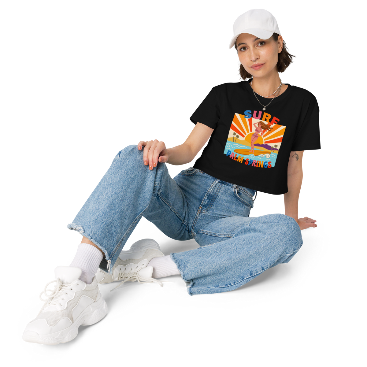 A woman wearing a black graphic t-shirt with a colorful retro surfboard design, light-wash wide-leg jeans, white socks, and white sneakers, sitting against a plain white background.