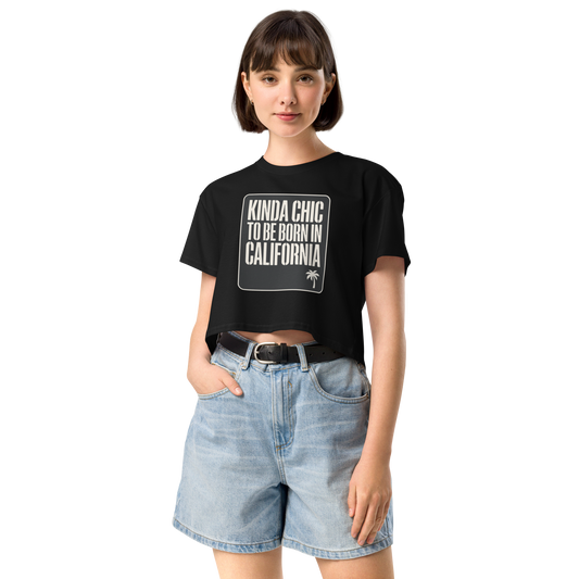 A woman modeling a black cropped t-shirt with a white graphic that reads 'KINDA CHIC TO BE BORN IN CALIFORNIA' alongside a small palm tree icon, paired with light-wash denim shorts against a plain white background.