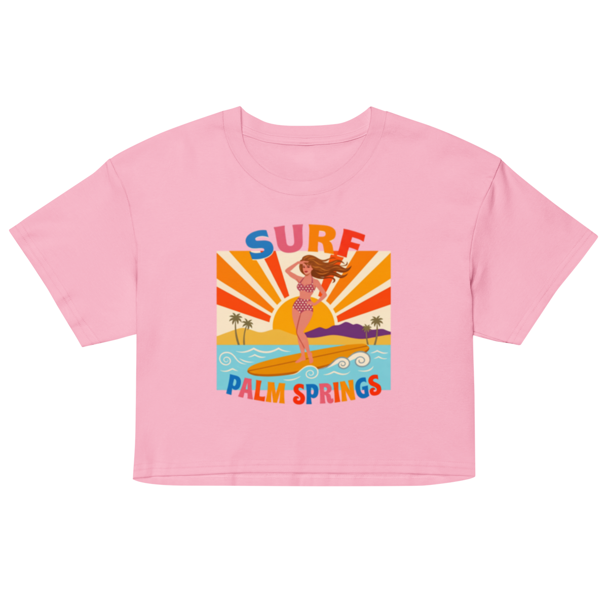 A light pink short-sleeve crop top t-shirt featuring a retro-style graphic of a woman surfing on a yellow board with palm trees and the text 'SURF PALM SPRINGS' in colorful letters.