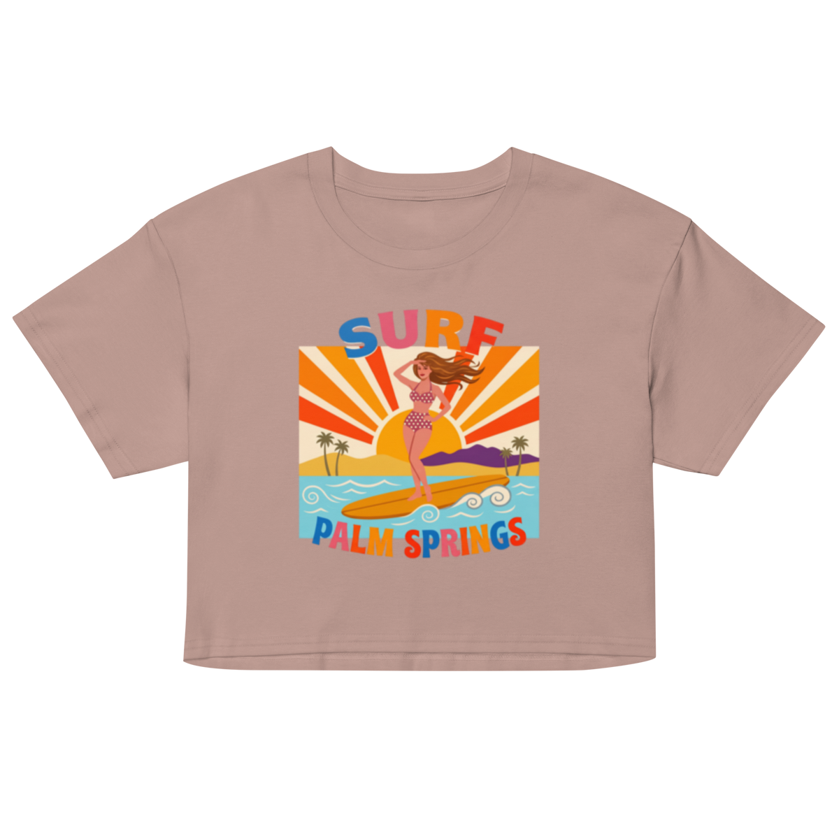 A cropped short-sleeve t-shirt in a dusty mauve-pink color, featuring a colorful retro graphic of a woman surfing on a surfboard with palm trees and the text 'SURF PALM SPRINGS'.