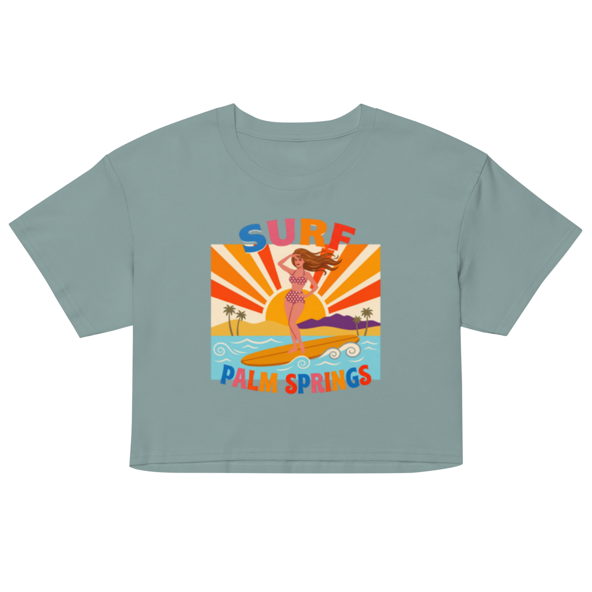 A sage green short-sleeve crop top featuring a colorful retro graphic of a woman surfing on a yellow board with palm trees and the text 'SURF PALM SPRINGS'.