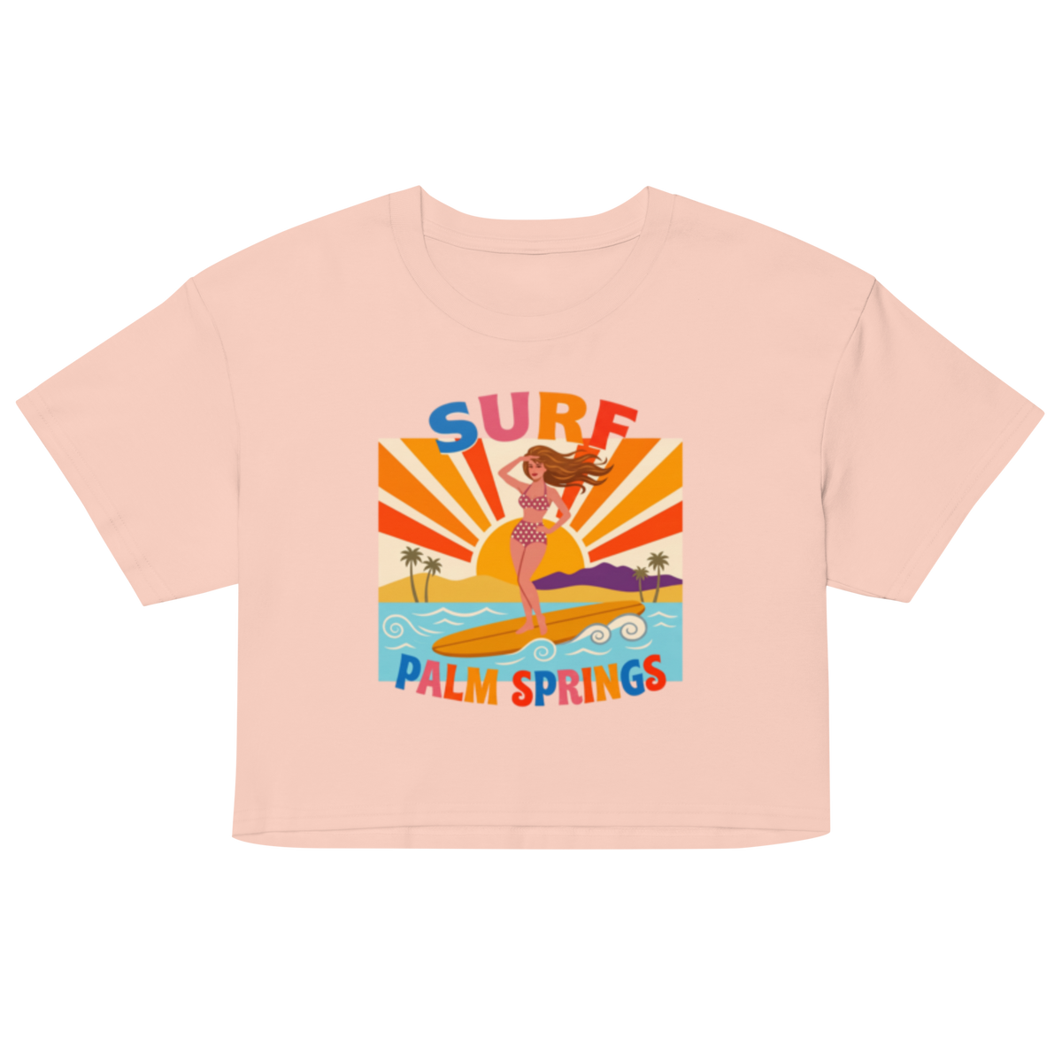 A light peach-pink short-sleeve crop top featuring a colorful retro graphic of a woman surfing on a yellow board with the text 'SURF' above and 'PALM SPRINGS' below, shown on a plain white background.