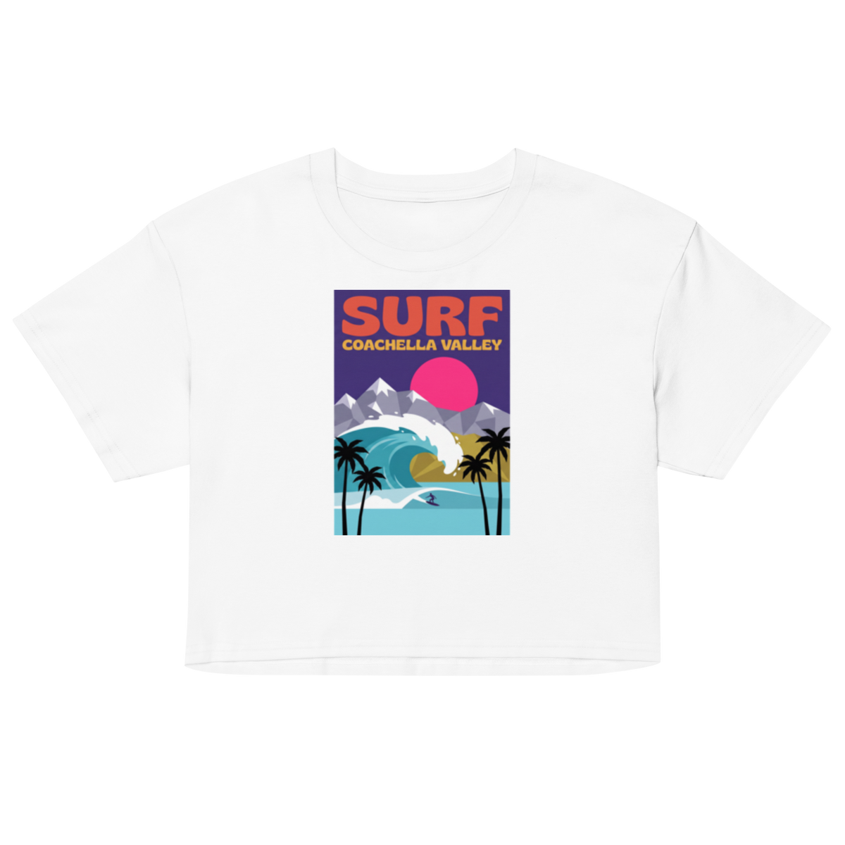 A white short-sleeve crop top t-shirt featuring a colorful retro-style graphic print on the front that reads 'SURF COACHELLA VALLEY' above an illustration of a surf wave, mountains, palm trees, and a pink sun.