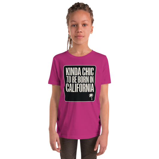 A young girl with braided hair wearing a magenta pink t-shirt with a black graphic box on the chest that reads 'KINDA CHIC TO BE BORN IN CALIFORNIA' in white text, standing against a plain white background.