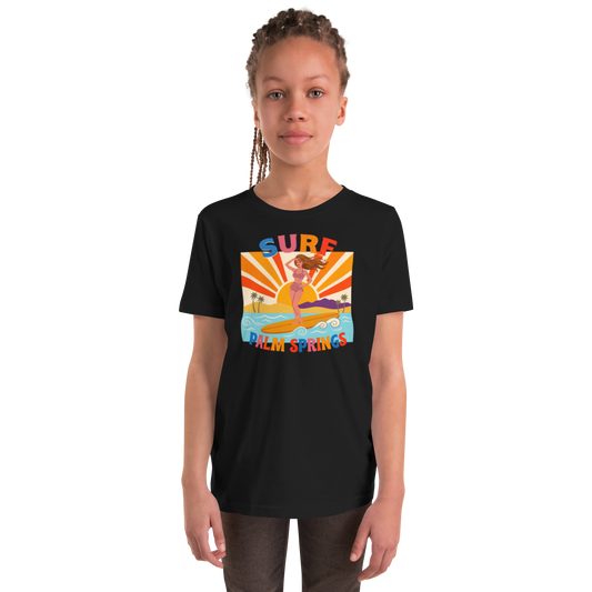 A young girl with braided hair wearing a black short-sleeve t-shirt with a colorful retro graphic that reads 'SURF Palm Springs' and shows a woman in a yellow bikini on a surfboard. The photo is shot against a clean white background.