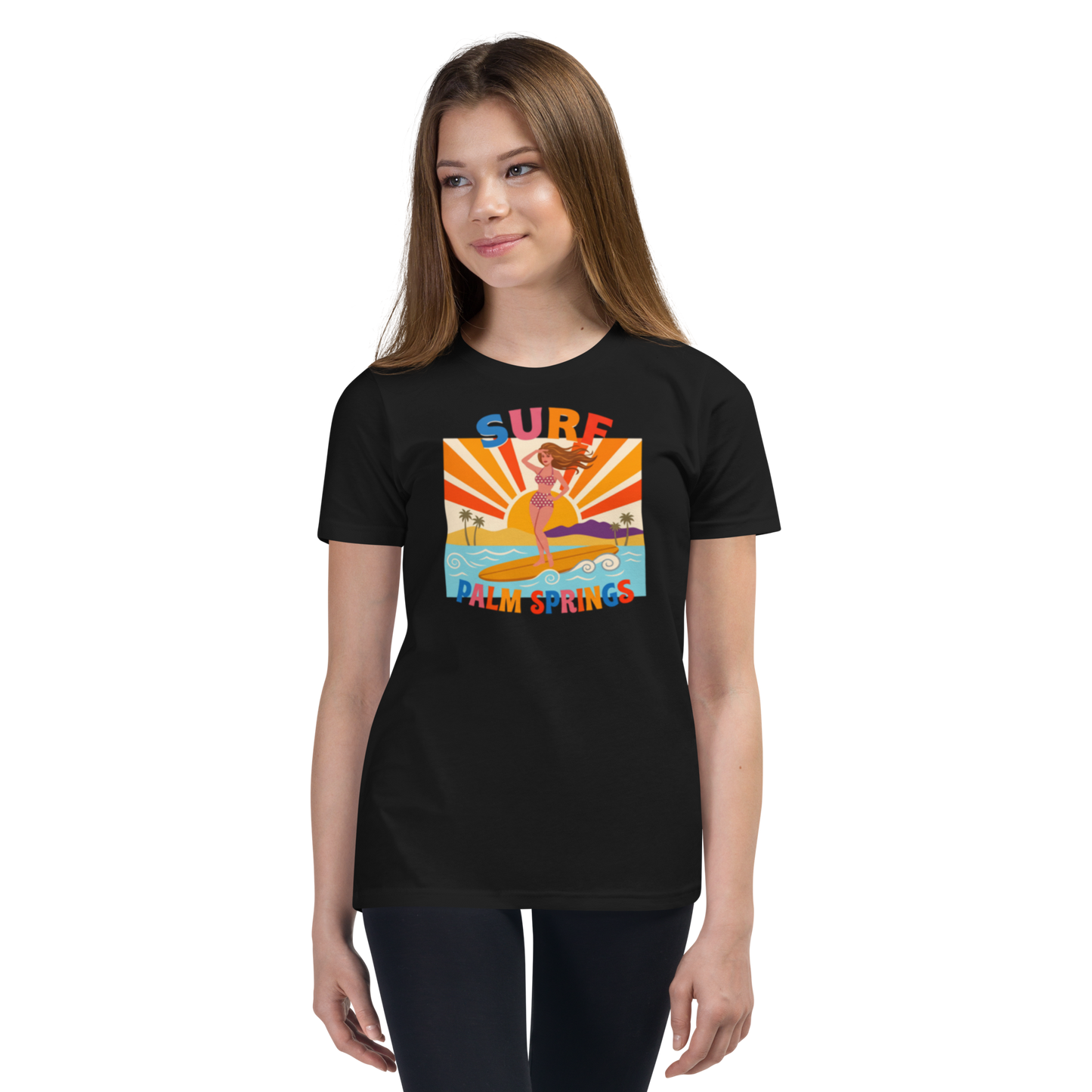 A young woman modeling a black short-sleeve t-shirt with a colorful retro-style graphic on the chest that reads 'SURF PALM SPRINGS' around an illustration of a surfer in a yellow surfboard. The background is a clean white studio backdrop.