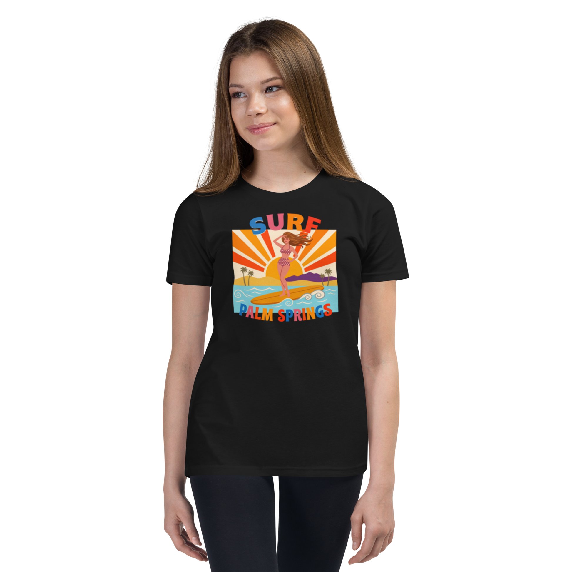 A young woman modeling a black short-sleeve t-shirt with a colorful retro-style graphic on the chest that reads 'SURF PALM SPRINGS' around an illustration of a surfer in a yellow surfboard. The background is a clean white studio backdrop.