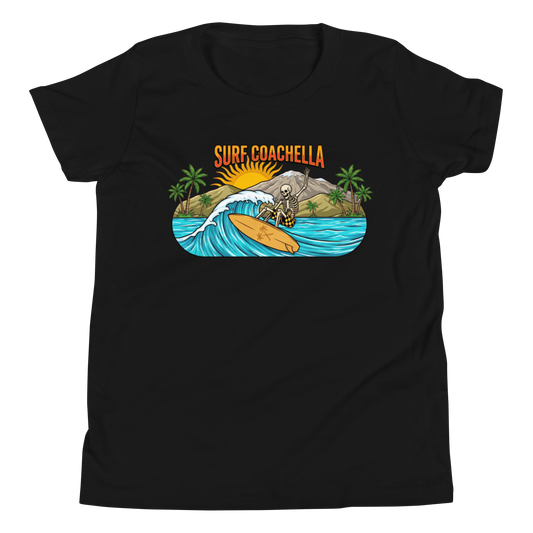 A black short-sleeve crewneck t-shirt featuring a colorful graphic print on the chest. The graphic shows a skeleton surfer riding an orange surfboard on a blue wave, with palm trees, a mountain, a rising sun, and the text 'SURF COACHELLA' in orange above.